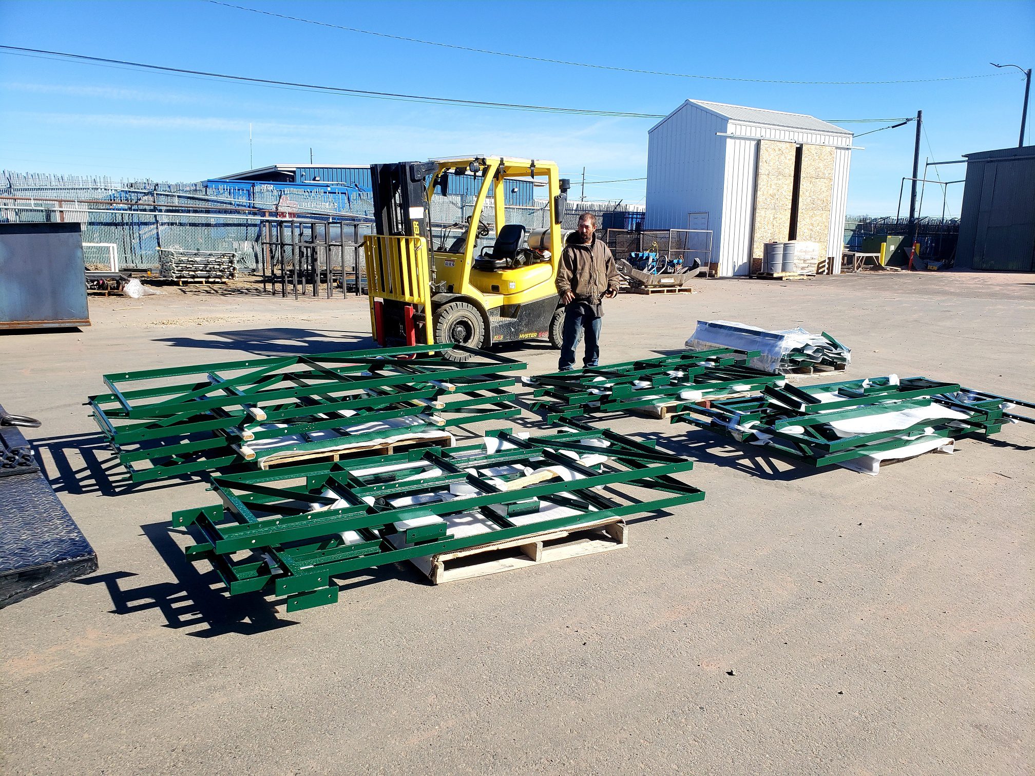 A worker stands near green metal frames and a yellow forklift outside.