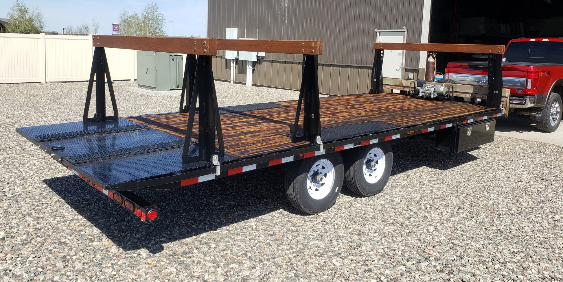 A flatbed trailer with dual axles and white wheels parked on gravel.